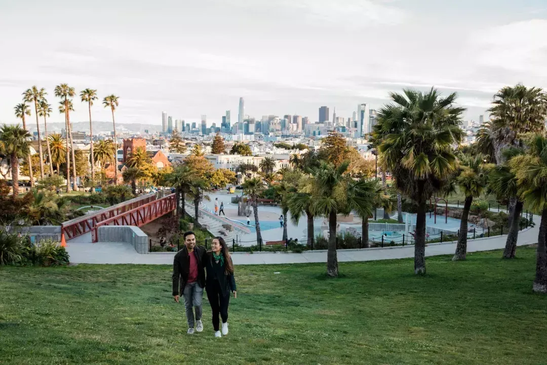 Una pareja camina hacia la cámara con Dolores Park y el Skyline 贝博体彩app detrás de ellos.